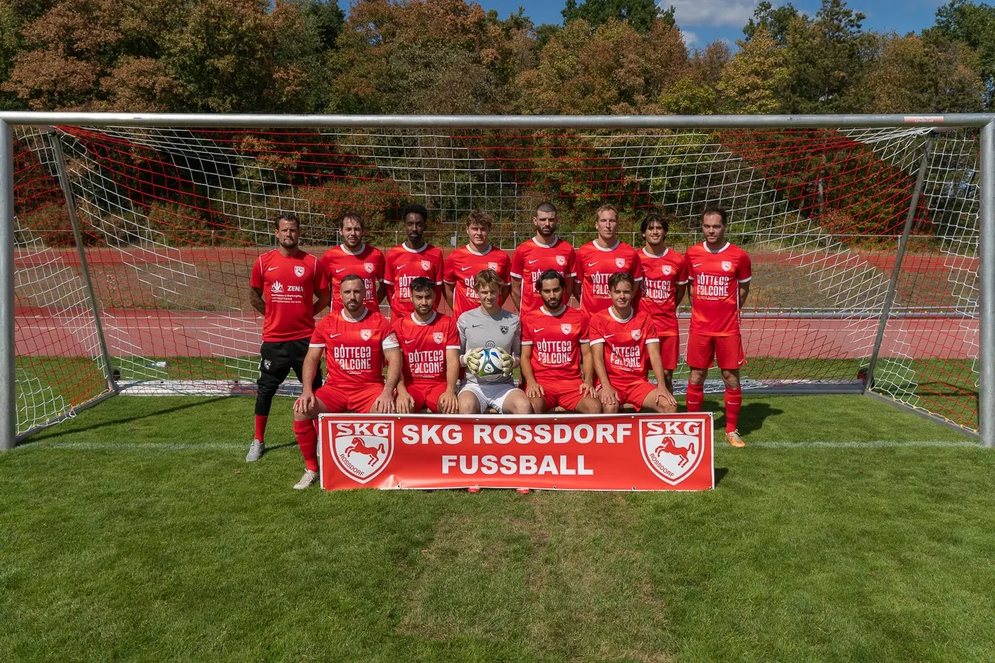 Die erste Fußballmannschaft der SKG Roßdorf in roten Trikots vor dem Tor mit rotem Netz, vorn ein Banner „SKG Rossdorf Fußball“, dahinter Herbstwald